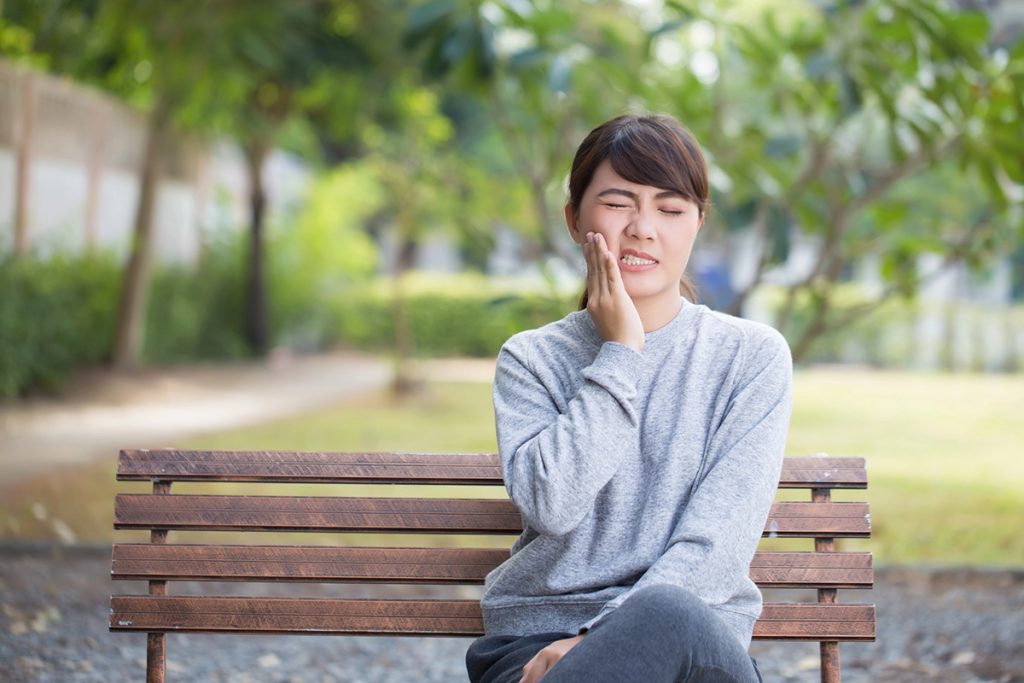 A woman sitting on a bench outdoors holding her cheek in pain, showing discomfort from a cavity.