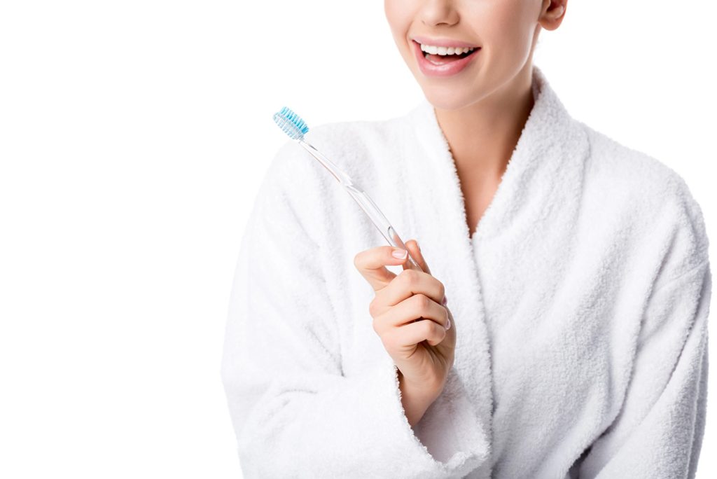 A woman in a white bathrobe smiling while holding a toothbrush with blue bristles against a plain white background.